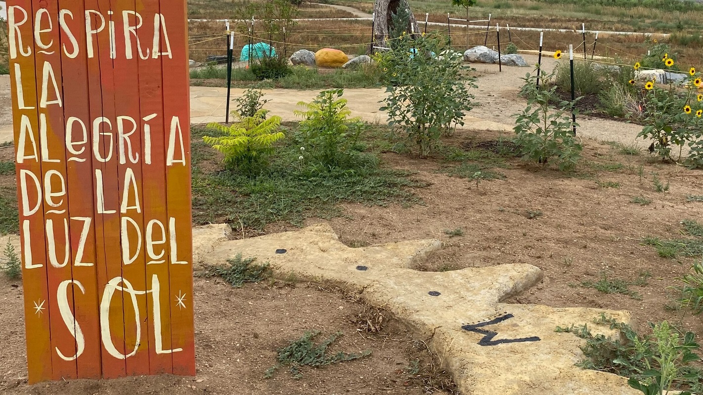 A wood sign painted in white Spanish text stands in the dirt with sunflowers and turquoise painted rocks