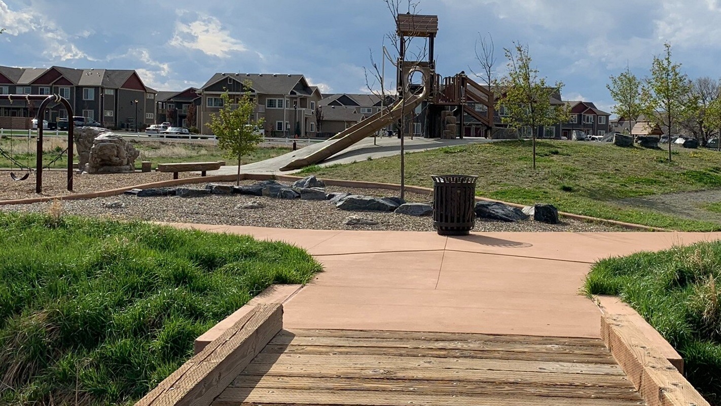 A wood slat path leads to a brown and tan playground with a slide, swings, green grass on the sides and apartments in the background